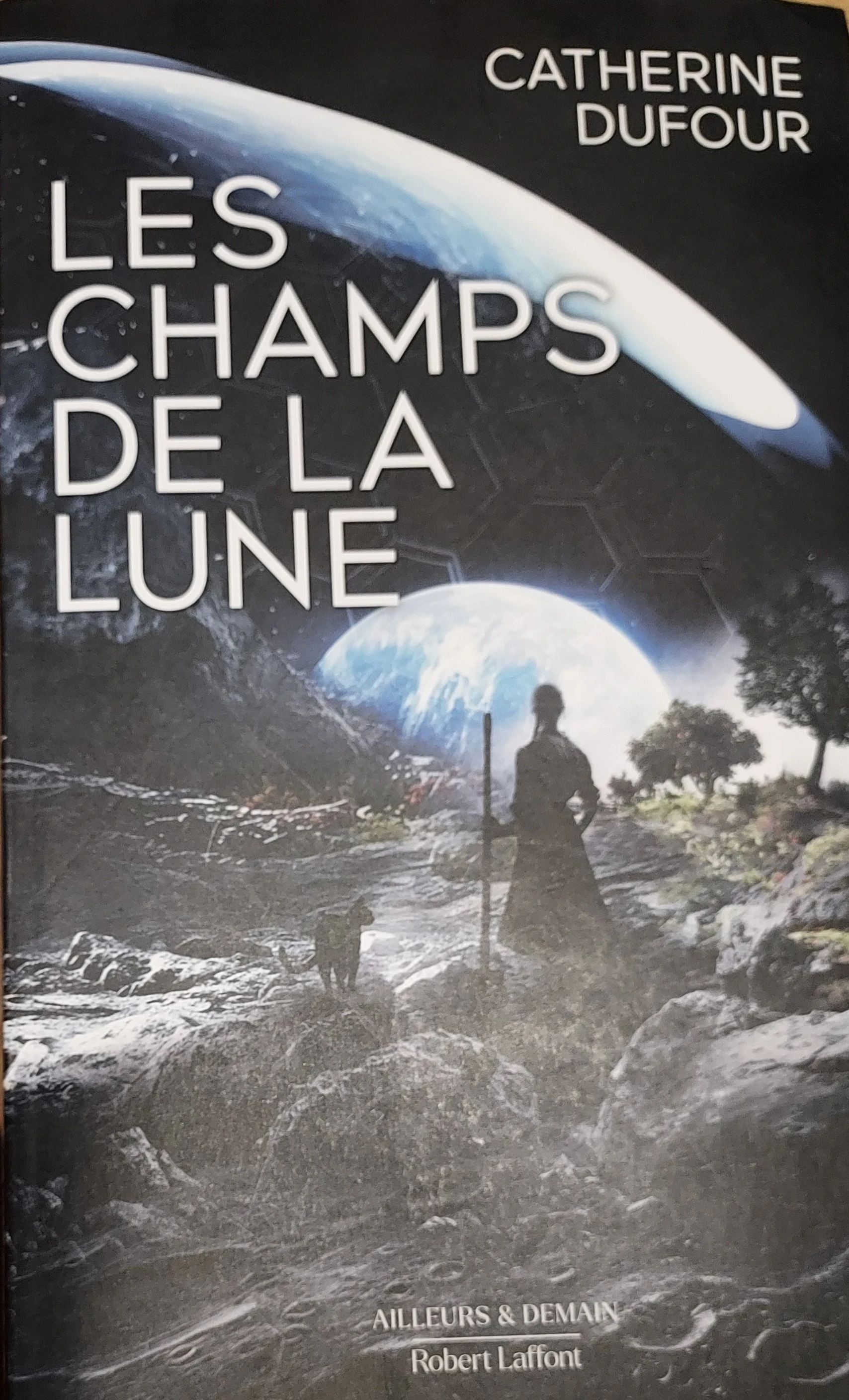 Illustration de Aurélien POLICE. Au premier plan, le sol lunaire, sombre, présente des cratères. Au centre, deux silhouettes, l'une féline, l'autre humaine tenant un bâton, nous tourne le dos et contemple un levé de Terre. Grosse sphère bleue apparaissant à l'horizon. Entre les deux, un peu de verdure, herbes et arbustes. Au-dessus des personnages, un reflet lumineux révèle la présence d'un dôme protecteur. Le titre et le nom de l’autrice sont en grosse lettres blanches.